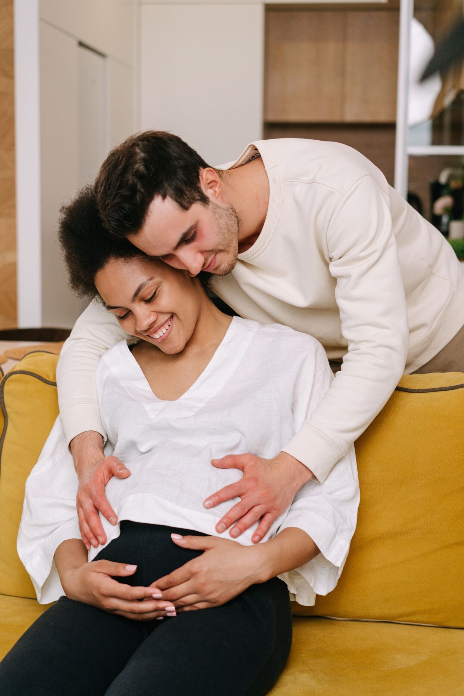 An expectant couple on a sofa at home, the partner gently hugging her from behind