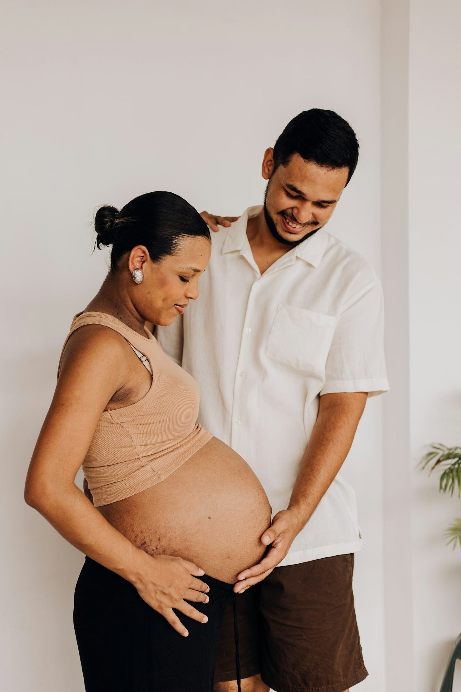 An expectant couple standing together, the partner smiling as she rests her hands on her bump